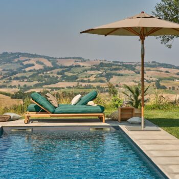 Zwei grüne, gepolsterte Liegestühle und ein beiger Sonnenschirm von Weishäupl aus Teakholz stehen am Pool und bieten unter strahlend blauem Himmel einen Blick auf sanfte Hügel und die umliegende Landschaft.