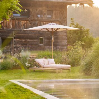 Ein gepolsterter Liegestuhl für den Außenbereich mit einem Weishäupl-Teakschirm steht auf dem Rasen neben einem Pool, eingerahmt von einem rustikalen Holzgebäude und üppigen Gartenpflanzen im Hintergrund.