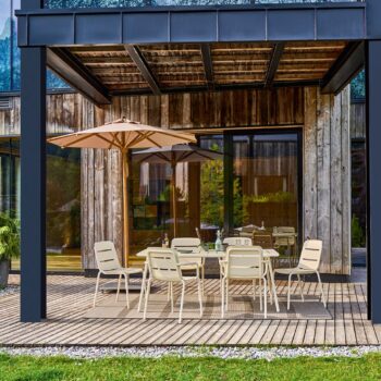 Eine moderne Terrasse mit Holzbelag, beigen Stühlen, Weishäupl-Tischen und einem großen Teakschirm vor einem Gebäude mit Glastüren und rustikaler Holzverkleidung.