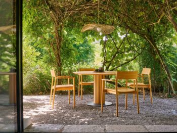 Ein Weishäupl-Tisch mit vier Stühlen steht auf Kies unter einer begrünten Pergola, während das Sonnenlicht durch dichtes grünes Laub in eine friedliche Gartenlandschaft fällt.