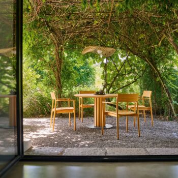 Ein Weishäupl-Tisch mit vier Stühlen steht auf Kies unter einer begrünten Pergola, während das Sonnenlicht durch dichtes grünes Laub in eine friedliche Gartenlandschaft fällt.