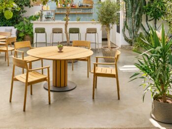 Sitzplatz im Freien mit rundem Weishäupl-Tisch und Stuhl, umgeben von üppiger Bepflanzung; im Hintergrund befindet sich eine Bar mit Hockern unter einem teilweise überdachten Dach.