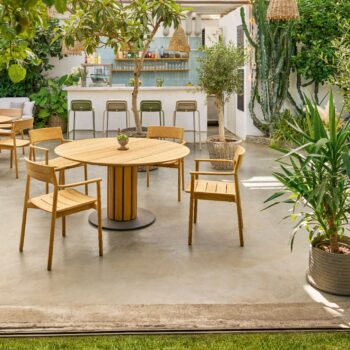 Sitzplatz im Freien mit rundem Weishäupl-Tisch und Stuhl, umgeben von üppiger Bepflanzung; im Hintergrund befindet sich eine Bar mit Hockern unter einem teilweise überdachten Dach.