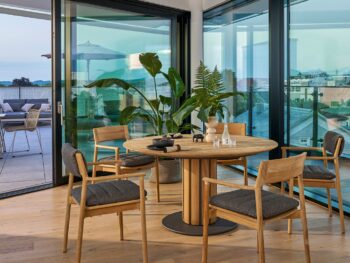 Ein moderner Essbereich mit rundem Weishäupl-Tisch, vier gepolsterten Stühlen, Topfpflanzen und großen Glastüren, die auf einen Balkon mit Sitzgelegenheiten im Freien führen.