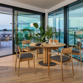 Ein moderner Essbereich mit rundem Weishäupl-Tisch, vier gepolsterten Stühlen, Topfpflanzen und großen Glastüren, die auf einen Balkon mit Sitzgelegenheiten im Freien führen.