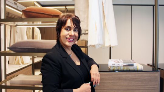 Eine Frau in einem schwarzen Blazer steht lächelnd neben einem Regal mit Kissen und Büchern in einem modernen, gut beleuchteten Innenraum.