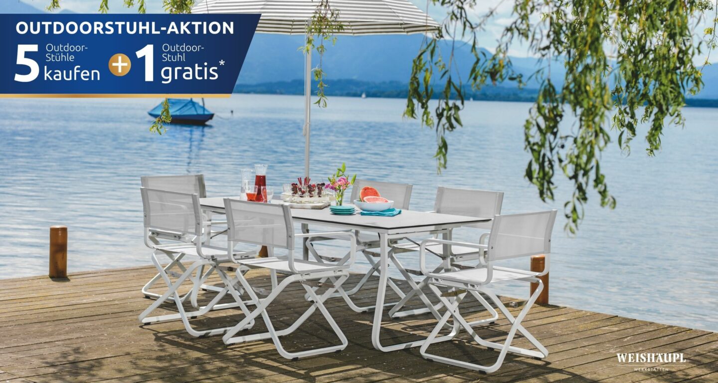 Eine weiße Outdoor-Essgarnitur vom Möbelhaus Bad Homburg mit sechs Stühlen steht auf einer Holzterrasse an einem See unter einem gestreiften Sonnenschirm, umgeben von Bäumen und Bergen.