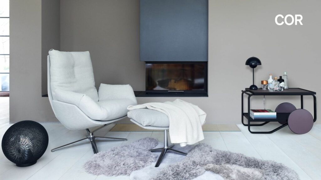 Modernes Wohnzimmer mit weißem Cordia Lounge Sessel und Ottomane, grauen Teppichen, schwarzem Kamin, Beistelltisch mit Dekor und dem Wort COR an der Wand - ein Raum, in dem Perfektion auf Komfort trifft.