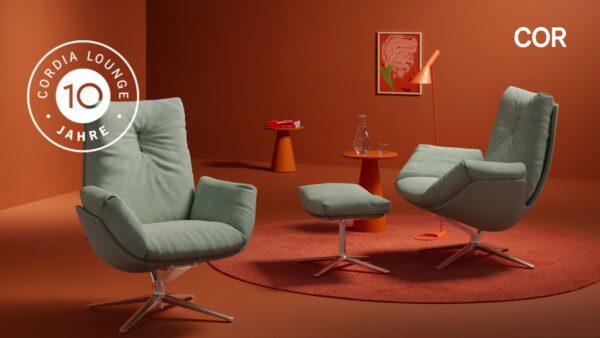 Zwei grüne Cordia Lounge Sessel mit passender Ottomane stehen auf einem orangefarbenen Teppich in einem modernen, orangefarbenen Raum und feiern Perfektion und Jubiläum mit 