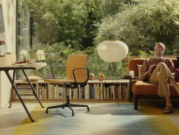 Ein Mann sitzt auf einem Sofa und liest auf einem Tablet in einem modernen Wohnzimmer mit großem Fenster, Schreibtisch, Bücherregal und Lampe - ein Beweis dafür, dass man schön wohnen und besser arbeiten kann, während man den Blick auf das üppige Grün draußen genießt.