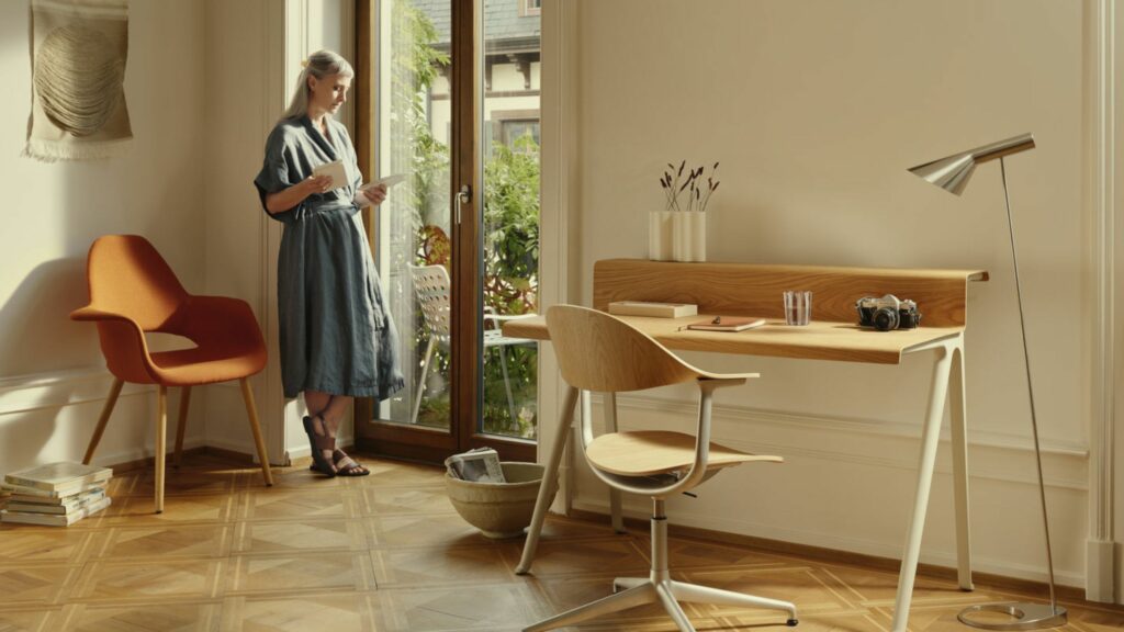 Eine Frau steht an einer Glastür und liest eine Zeitschrift in einem hellen Raum mit Parkettboden, Holzschreibtisch, orangefarbenem Sessel und Stehlampe - ein inspirierendes Homeoffice für schön wohnen und besser arbeiten.
