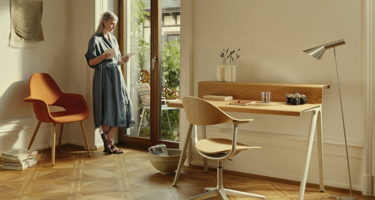 Eine Frau steht an einer Glastür und liest eine Zeitschrift in einem hellen Raum mit Parkettboden, Holzschreibtisch, orangefarbenem Sessel und Stehlampe - ein inspirierendes Homeoffice für schön wohnen und besser arbeiten.