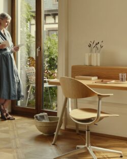 Eine Frau steht an einer Glastür und liest eine Zeitschrift in einem hellen Raum mit Parkettboden, Holzschreibtisch, orangefarbenem Sessel und Stehlampe - ein inspirierendes Homeoffice für schön wohnen und besser arbeiten.