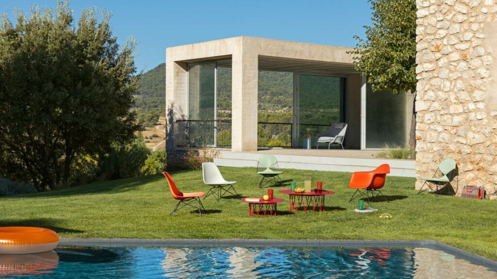 Moderne Terrasse mit farbenfrohen Vitra Stühlen und Tischen, echte Designklassiker, angeordnet auf Gras in der Nähe eines Steinhauses, mit einem Pool im Vordergrund und Hügeln im Hintergrund - exklusiv kuratiert von Möbel Braum.