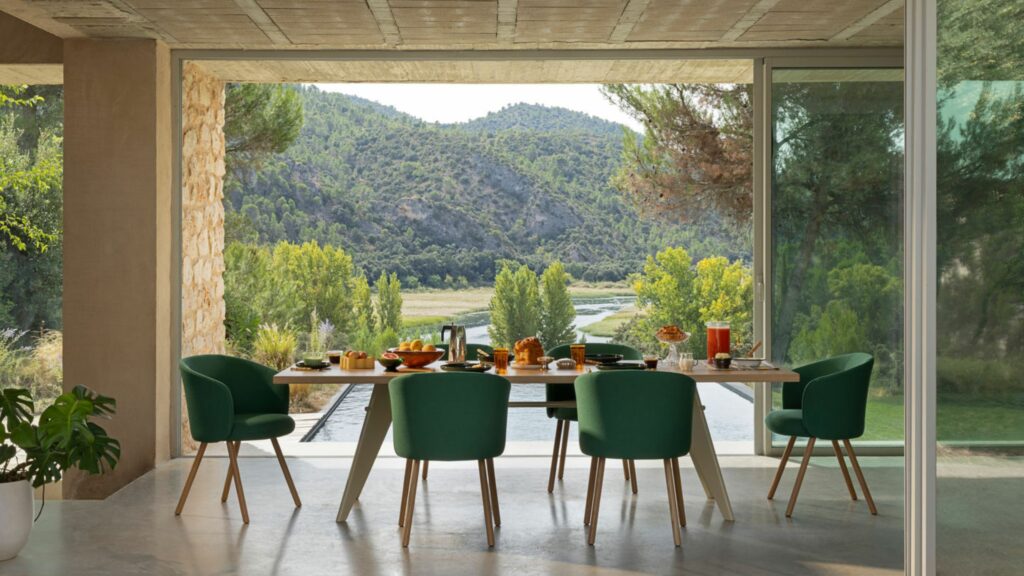 Ein Esstisch mit sechs grünen Vitra-Stühlen steht vor großen Glastüren, die den Blick auf eine malerische Landschaft mit Bäumen, Hügeln und einem Fluss freigeben - ein einladender Raum, der von Möbel Braum kuratiert wurde.