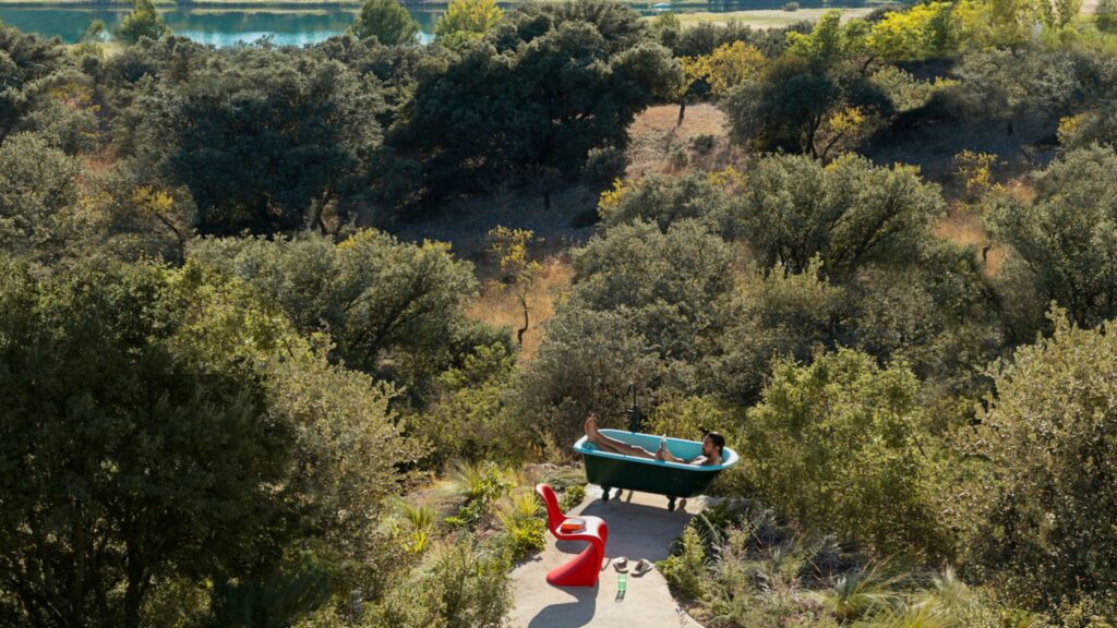 Eine Person entspannt sich in einer freistehenden Badewanne im Freien auf einem Steinweg, mit zwei roten Vitra Designklassiker-Stühlen in der Nähe und umgeben von dichten Bäumen und Grün.
