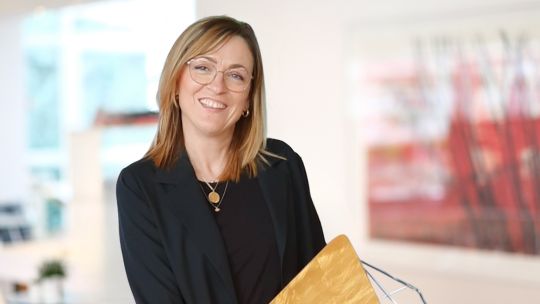 Eine Frau mit schulterlangem Haar, Brille und schwarzem Blazer hält einen goldfarbenen Ordner in der Hand und steht in einem modernen, hell erleuchteten Büroraum.