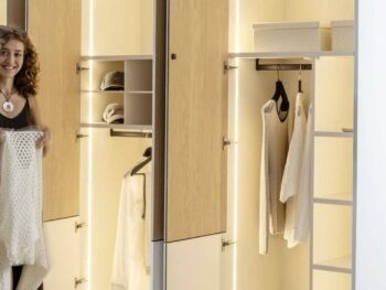 Sudbrock Möbel: Eine Frau steht vor einem offenen, gut beleuchteten Kleiderschrank mit einem weißen Pullover in der Hand, in dem Kleidung und Aufbewahrungsboxen organisiert sind.