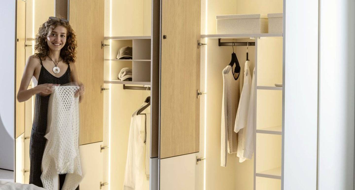 Sudbrock Möbel: Eine Frau steht vor einem offenen, gut beleuchteten Kleiderschrank mit einem weißen Pullover in der Hand, in dem Kleidung und Aufbewahrungsboxen organisiert sind.