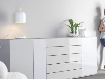 Sudbrock Möbel: Eine Frau steht an einem modernen weißen Sideboard mit Schubladen, auf dem Zeitschriften liegen. Auf der Anrichte befinden sich eine Lampe, eine Pflanze und Dekorationsgegenstände. Im Vordergrund steht ein weißer Stuhl.