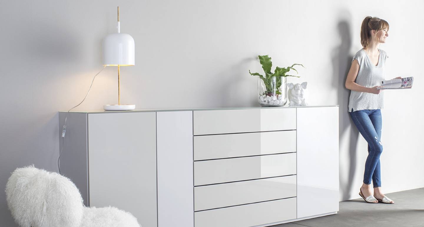 Sudbrock Möbel: Eine Frau steht an einem modernen weißen Sideboard mit Schubladen, auf dem Zeitschriften liegen. Auf der Anrichte befinden sich eine Lampe, eine Pflanze und Dekorationsgegenstände. Im Vordergrund steht ein weißer Stuhl.