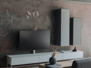 Modernes Wohnzimmer mit einer Team 7 cubus pure Wohnwand, mit einem Flachbildfernseher auf einem niedrigen weißen Schrank, zwei hohen Hängeschränken, einem grauen Sofa und einem minimalistischen Couchtisch mit einer Vase und Trockenblumen.
