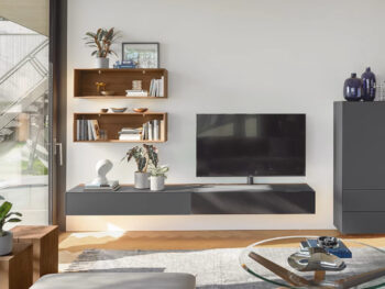 Modernes Wohnzimmer mit einer reinen Wohnwand von Team 7 cubus mit wandmontiertem Fernseher, schwebendem Schrank, Holzregalen mit Büchern und Pflanzen, Couchtisch aus Glas und dekorativen Vasen. Ein großes Fenster erfüllt den Raum mit natürlichem Licht.