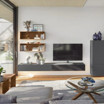 Modernes Wohnzimmer mit einer reinen Wohnwand von Team 7 cubus mit wandmontiertem Fernseher, schwebendem Schrank, Holzregalen mit Büchern und Pflanzen, Couchtisch aus Glas und dekorativen Vasen. Ein großes Fenster erfüllt den Raum mit natürlichem Licht.