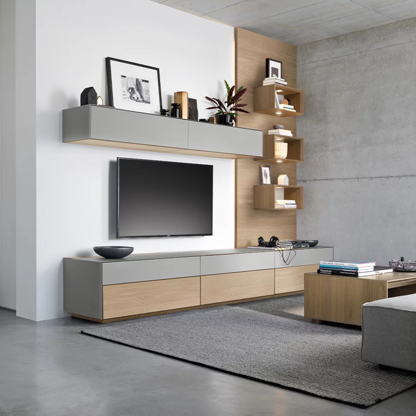 Modernes Wohnzimmer mit einer Team 7 cubus pure Wohnwand, wandmontiertem TV, minimalistischen hölzernen und grauen Schränken, schwebenden Regalen mit Dekor und einem grauen Teppich auf Betonboden. Modernes Wohnzimmer mit einer Team 7 cubus pure Wohnwand, wandmontiertem TV, minimalistischen hölzernen und grauen Schränken, schwebenden Regalen mit Dekor und einem grauen Teppich auf Betonboden.