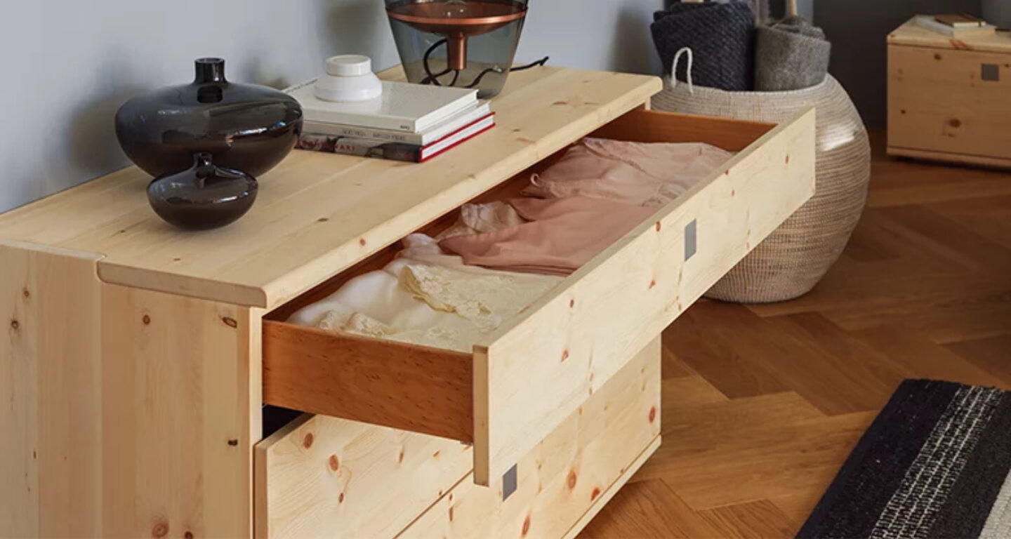 Eine Kommode aus hellem Holz mit offenen Schubladen, in denen sich ordentlich gefaltete Kleidung befindet; oben auf der Kommode befinden sich Dekorationsartikel und Bücher.