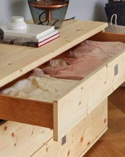 Eine Kommode aus hellem Holz mit offenen Schubladen, in denen sich ordentlich gefaltete Kleidung befindet; oben auf der Kommode befinden sich Dekorationsartikel und Bücher.
