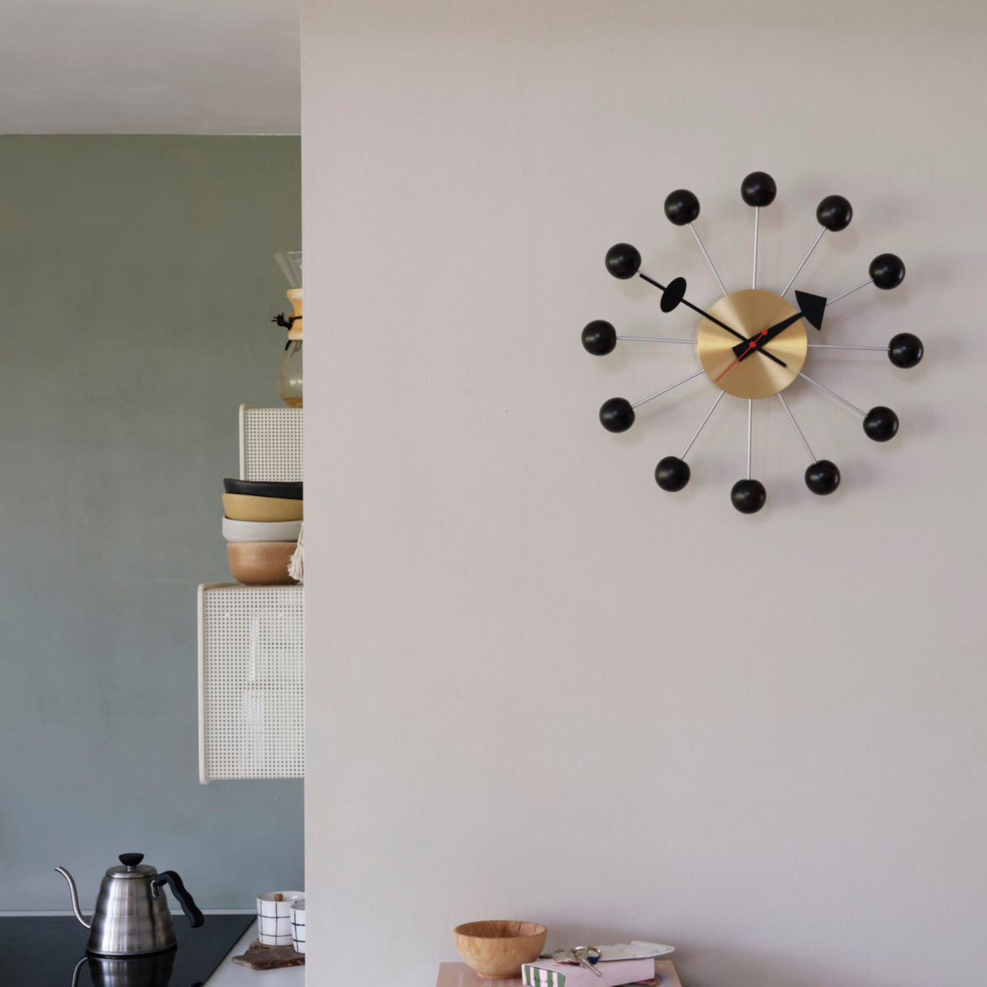 Eine Vitra Ball Clock mit schwarzen Kugeln ist an einer weißen Wand montiert; im Hintergrund sind Küchenutensilien und ein Wasserkocher zu sehen, die dem Raum einen Hauch von Designer-Uhr-Stil verleihen.