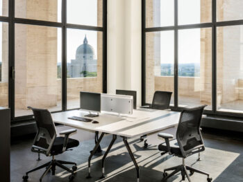 Moderner Büroraum mit vier Schreibtischen und Stühlen, die um zwei Computermonitore herum angeordnet sind, in der Nähe großer Fenster mit Blick auf ein Kuppelgebäude.