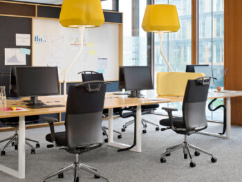 Moderner Büroarbeitsplatz mit mehreren Schreibtischen, schwarzen Drehstühlen, Computern, gelben Lampen und großen Fenstern, die natürliches Licht hereinlassen.