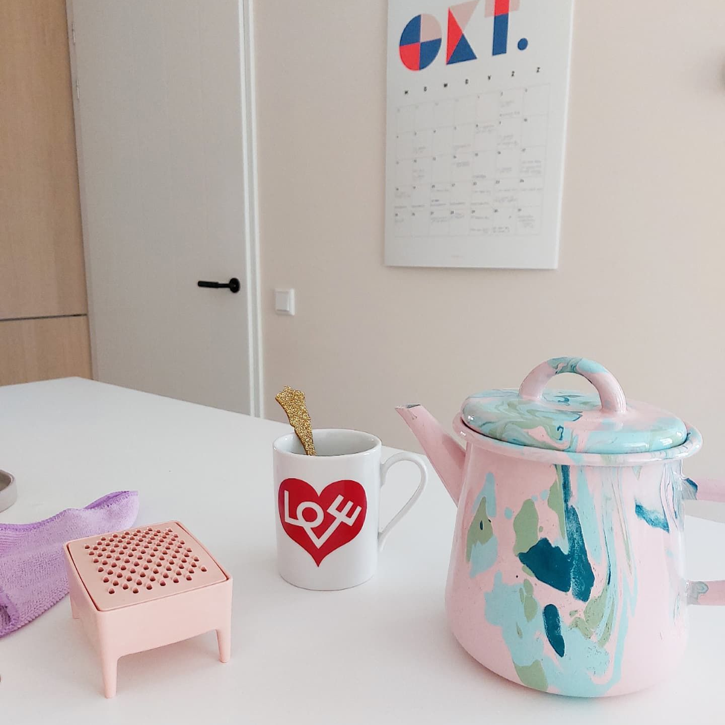 Eine Teekanne aus pastellfarbenem Marmor, ein Vitra-Kaffeebecher mit rotem LOVE-Motiv, eine rosa Reibe und ein Putztuch stehen auf einer weißen Arbeitsplatte; im Hintergrund hängt ein Kalender an der Wand.