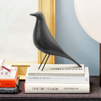 Eine schwarze Eames-House-Bird-Skulptur steht auf einem Bücherstapel neben einem goldgerahmten Bild und einer weißen Lampe und fügt der Szene das ikonische Vitra-Design hinzu.