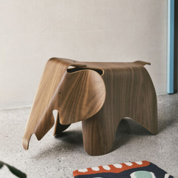 Hölzerner Vitra Elephant Hocker auf einem Betonboden neben einem bunt gemusterten Teppich und einer Wand.