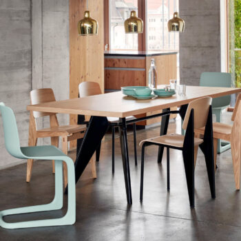 Moderner Essbereich mit einem EM-Tisch von Vitra, Stühlen aus gemischtem Holz und hellblauen Stühlen, Pendelleuchten und einem Geschirr-Set, alles in einem hellen Raum mit Betonwänden und großen Fenstern.