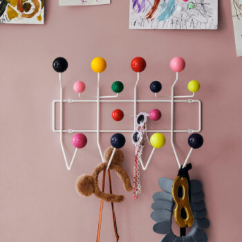 Eine weiße Vitra Hang it all Wandgarderobe mit bunten runden Haken hält ein Bärenohr-Stirnband, eine Perlenkette, eine Sonnenbrille und eine graue Maske vor einer rosa Wand mit Kinderzeichnungen.