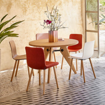 Ein runder Holztisch mit einer Kupfervase und Blumen ist von vier Stühlen umgeben - zwei rote, ein weißer Vitra Stuhl und ein Holzstuhl von Hal Wood - in einem sonnendurchfluteten Raum mit gemustertem Fliesenboden.