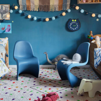 Ein Kinderzimmer mit zwei blauen Panton Chair Juniors von Vitra, einer Wanduhr, Dinosaurierspielzeug, Stofftieren, Lichterketten und buntem Dekor auf einem gemusterten Teppich.