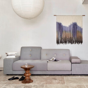 Ein modernes Wohnzimmer mit einem grauen Vitra-Sofa, einem kleinen Beistelltisch aus Holz, einer runden Hängeleuchte, gestapelten Büchern, Hausschuhen und einem gewebten Wandbehang mit Fransen.