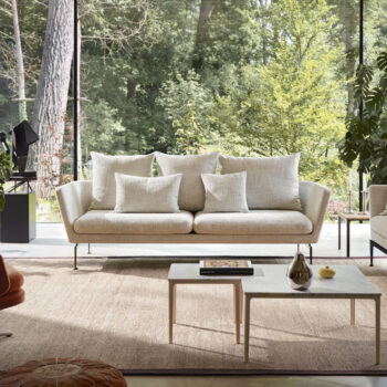 Ein modernes Wohnzimmer mit einem beigen Vitra Suita Sofa, mehreren weißen Kissen, zwei verschachtelten Couchtischen, einem Teppich und großen Fenstern mit Blick auf einen grünen Wald.