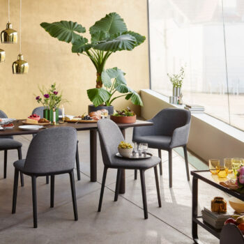 Ein moderner Essbereich mit Vitra-Softshell-Stühlen, einem Holztisch mit Speisen und Getränken, einer großen Topfpflanze und Dekorationsartikeln an einem bodentiefen Fenster.