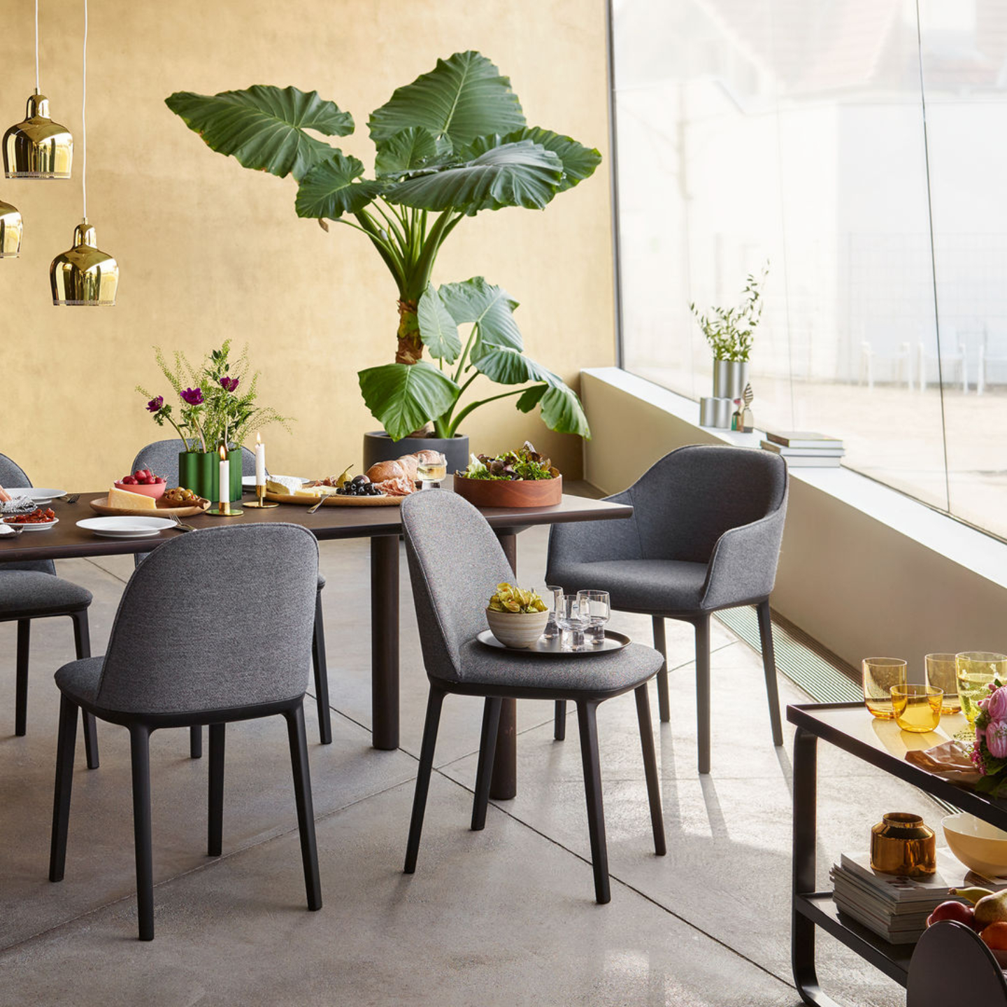 Ein moderner Essbereich mit Vitra-Softshell-Stühlen, einem Holztisch mit Speisen und Getränken, einer großen Topfpflanze und Dekorationsartikeln an einem bodentiefen Fenster.