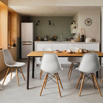 Moderne Küche und Essbereich mit einem Holztisch, vier weißen Stühlen, einer Kücheninsel, einem Kühlschrank und verschiedenen Utensilien auf den Arbeitsflächen. Durch ein großes Fenster fällt natürliches Licht ein.