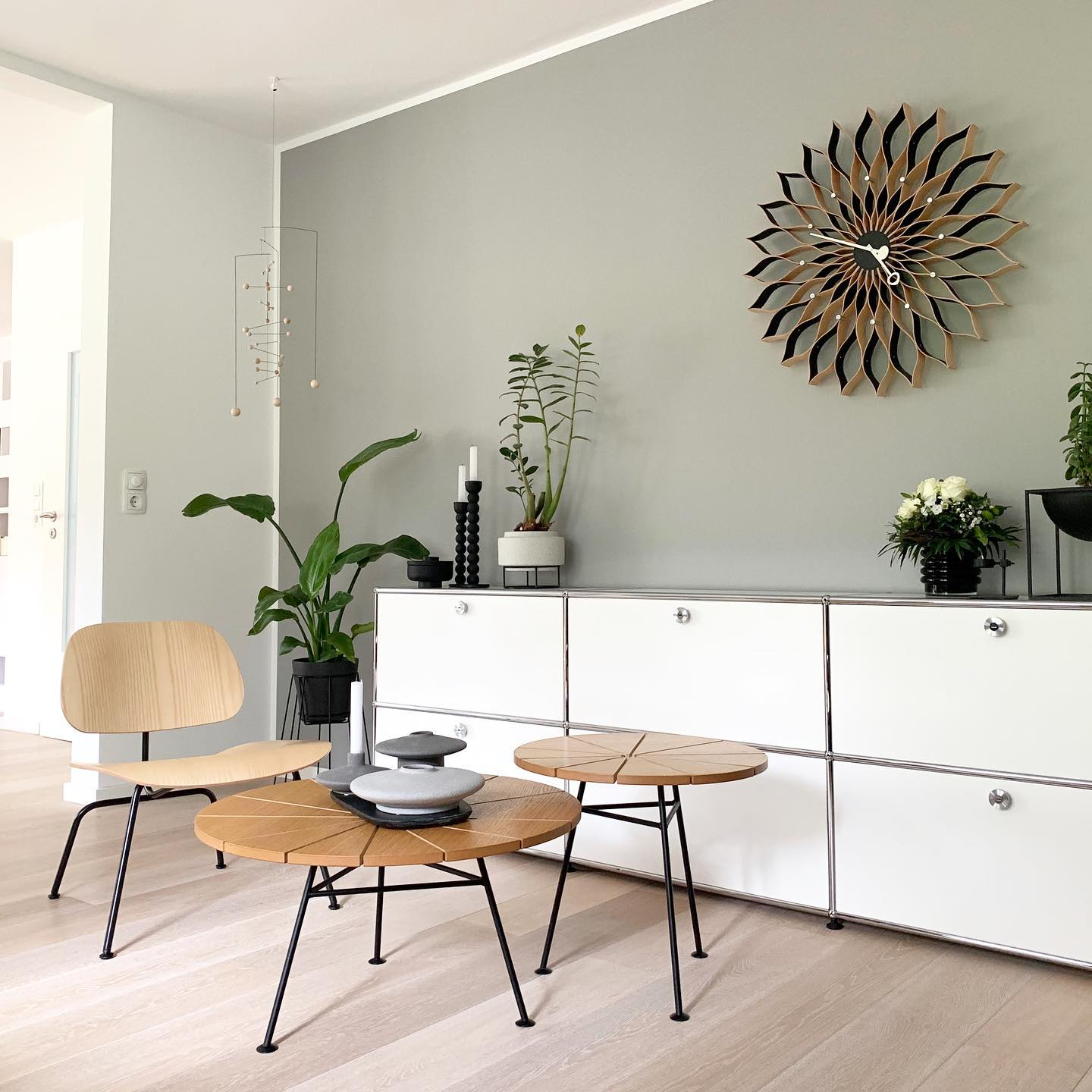 Modernes Wohnzimmer mit hellem Holzfußboden, minimalistischen Möbeln, Zimmerpflanzen und einer Vitra Sunflower Wanduhr über einem weißen Schrank.