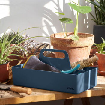 Eine blaue Vitra Toolbox mit Gartengeräten steht auf einem Holztisch, umgeben von Topfpflanzen und Erde, in einem sonnendurchfluteten Raum.