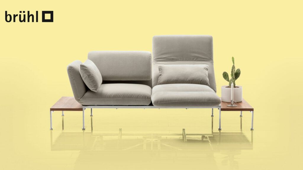 Modernes, hellgraues Modulsofa aus dem Brühl-Sofaprogramm „roro“ mit verstellbaren Rückenlehnen auf einem Gestell aus Holz und Metall, daneben ein kleiner Tisch mit einem Kaktus im Topf, vor einem gelben Hintergrund mit dem Brühl-Logo.