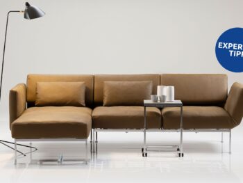 Ein modernes, braunes Sofaprogramm „roro“ von Brühl mit einer Chaiselongue, einem Beistelltisch aus Metall, auf dem Tassen gestapelt sind, und einer schwarzen Stehlampe vor einem schlichten weißen Hintergrund. An der Wand befindet sich ein blauer „Expertentipp“-Aufkleber.